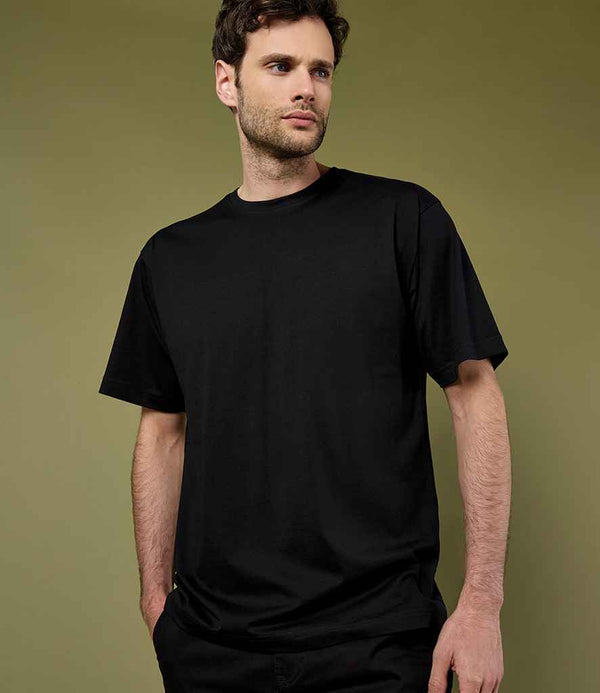 A man with short brown hair and a beard wears the Kustom Kit Hunky® Superior T-Shirt by Kustom Kit, featuring reinforced shoulder seams, along with black pants. He stands against an olive green background, looking slightly to his left.
