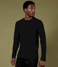 A man stands against an olive green background, wearing a Kustom Kit Long Sleeve Fashion Fit Superwash® 60°C T-Shirt and black pants. He looks to his left with a neutral expression.