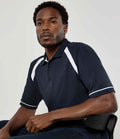 A man with short curly hair sits on a black chair, wearing the Kustom Kit Oak Hill Cotton Piqué Polo Shirt by Kustom Kit, which features contrast panels, white stripes, and a ribbed knit collar against a plain light background.