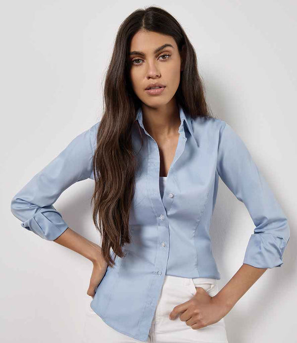 A woman with long brown hair wears the Kustom Kit Ladies Premium Long Sleeve Tailored Oxford Shirt by Kustom Kit in light blue, paired with white pants. She stands against a plain wall, hands on hips, looking directly at the camera.