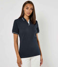 A woman with straight brown hair wears the Kustom Kit Ladies St Mellion Tipped Cotton Piqué Polo Shirt, a navy blue polo featuring a ribbed collar and pearl button placket by Kustom Kit, paired with white pants against a light background.