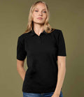 A woman with long blonde hair wears the Kustom Kit Ladies Regular Fit Workforce Piqué Polo Shirt by Kustom Kit, styled with blue jeans. She stands before an olive green background, looking directly at the camera with a neutral expression.