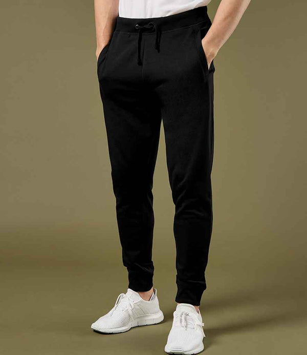 Someone wearing Kustom Kit Slim Fit Sweat Pants with an elasticated waistband, a white t-shirt, and white sneakers stands with hands in pockets against a plain olive green background.