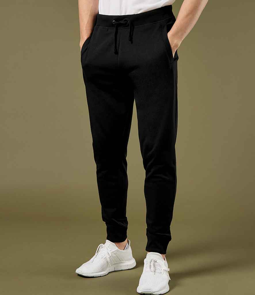 Someone wearing Kustom Kit Slim Fit Sweat Pants with an elasticated waistband, a white t-shirt, and white sneakers stands with hands in pockets against a plain olive green background.