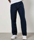 A person in Kustom Kit Cooltex® Track Pants (navy, straight-leg, elastic waistband) from Kustom Kit, a white t-shirt, and white sneakers stands against a neutral background with their hands behind their back and head out of frame.