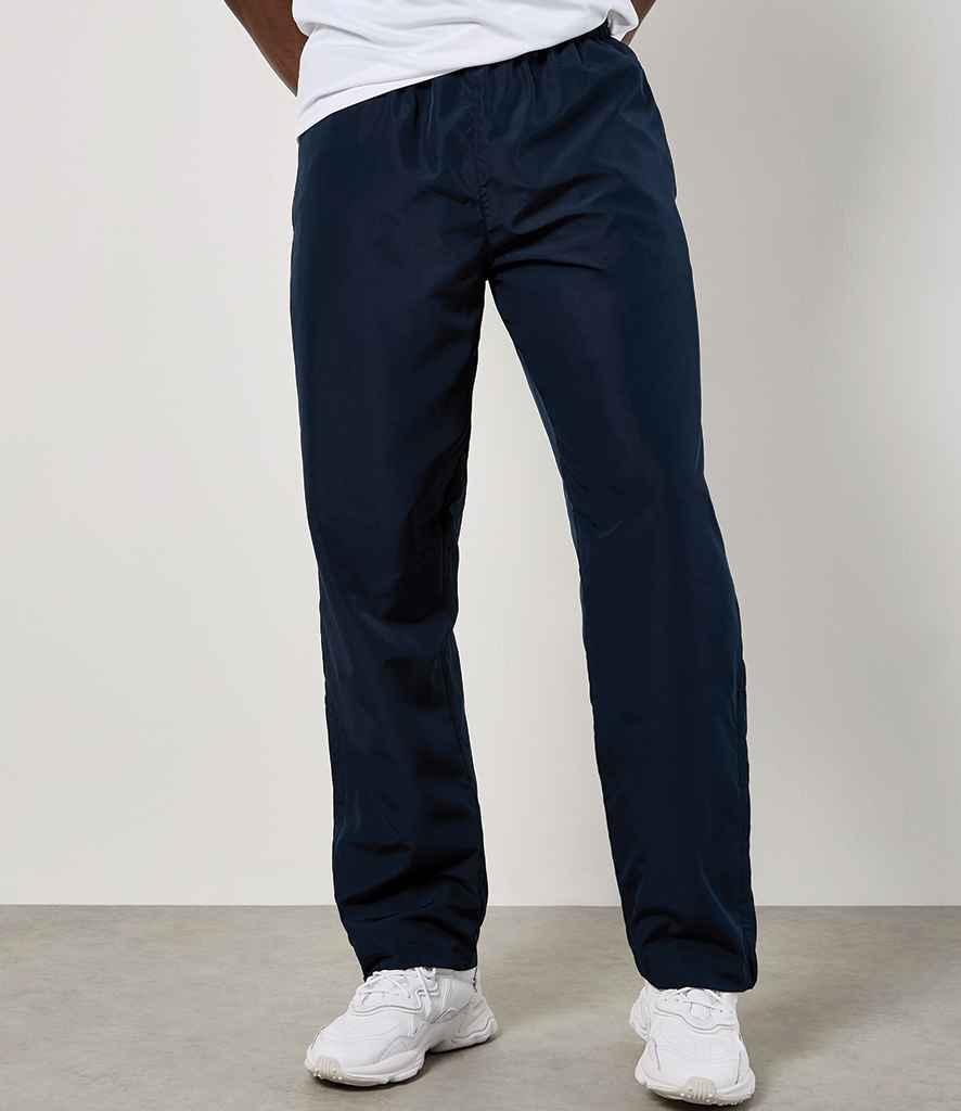 A person in Kustom Kit Cooltex® Track Pants (navy, straight-leg, elastic waistband) from Kustom Kit, a white t-shirt, and white sneakers stands against a neutral background with their hands behind their back and head out of frame.