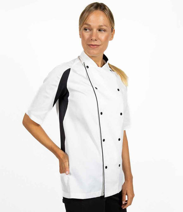 A person stands against a white background wearing the Le Chef Short Sleeve Executive Jacket by Le Chef, a unisex white double-breasted chef jacket with black side panels and buttons. Their right hand is in their pocket and they are looking to their left.