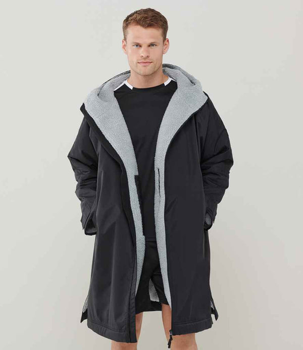 A man stands against a plain light background wearing the Finden + Hales Adults All Weather Robe with a light gray sherpa fleece lining over a black t-shirt and shorts, looking at the camera with his hands in his robe pockets.