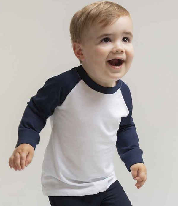 A young child with light hair smiles in a Larkwood Baby/Toddler Long Sleeve Baseball T-Shirt, featuring a white body with contrast collar and dark sleeves, paired with dark pants, against a plain light background.