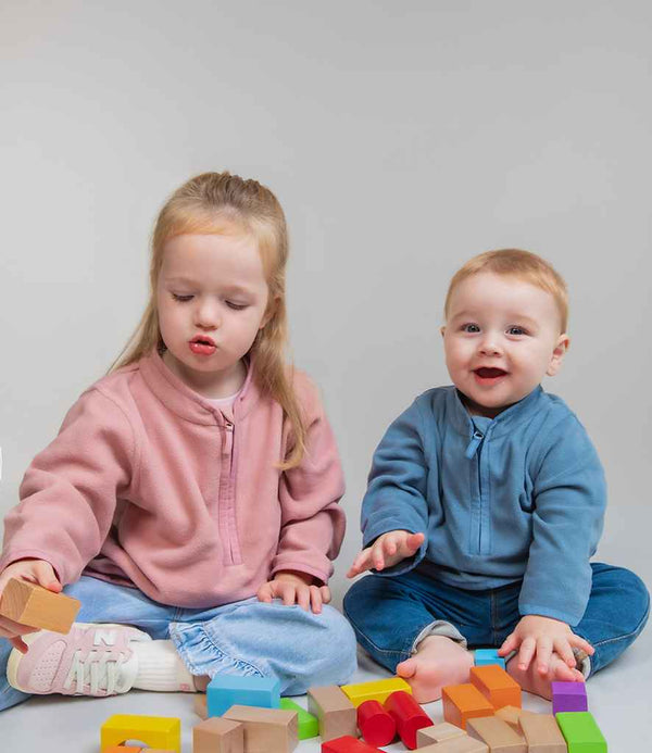 Two young children play on the floor with colorful wooden blocks. The boy on the right wears a blue Larkwood Kids 1/2 Zip Lightweight Fleece by Larkwood, paired with light pants, as both look toward the camera.