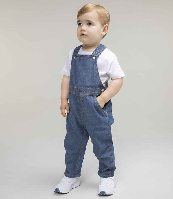 A young child with light hair stands slightly left, hands in pockets, wearing a white t-shirt, Larkwood Kids Denim Dungarees by Larkwood with rear patch pockets and jean stitching, plus white sneakers, against a plain background.