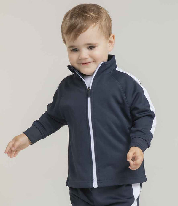 A toddler with light brown hair wears the Larkwood Baby/Toddler Tracksuit Top by Larkwood, a navy blue zip-up with white sleeve stripes, standing against a plain light background and looking down with a slight smile.