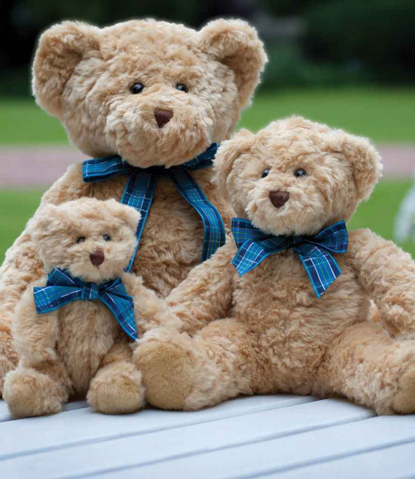 Three Mumbles Bracken Bears by Mumbles, each light brown and bean-filled with a blue tartan ribbon, sit together on a white surface against a blurred green outdoor backdrop.