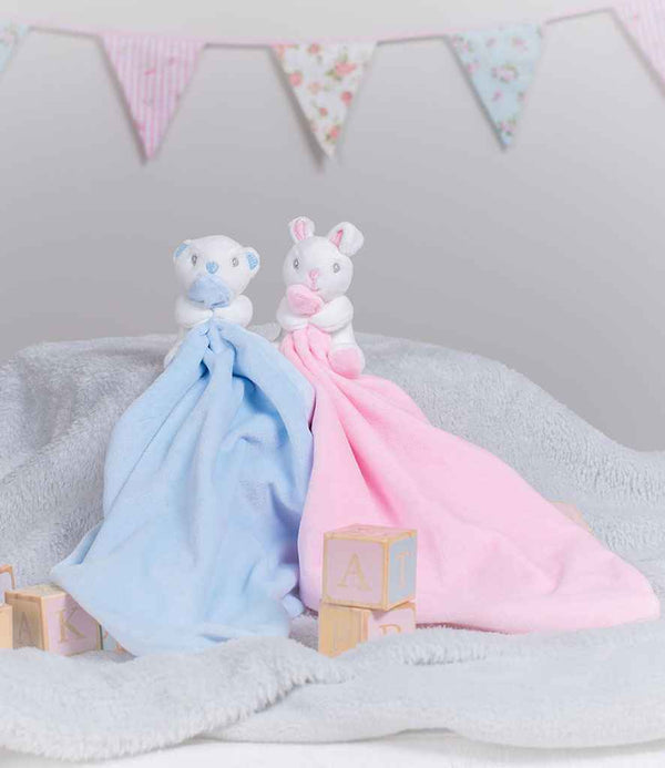 Two Mumbles Animal Comforter with Rattle toys—a bear with a blue blanket and a rabbit with a pink blanket—rest on a gray plush surface, surrounded by wooden letter blocks and pastel bunting in the background.