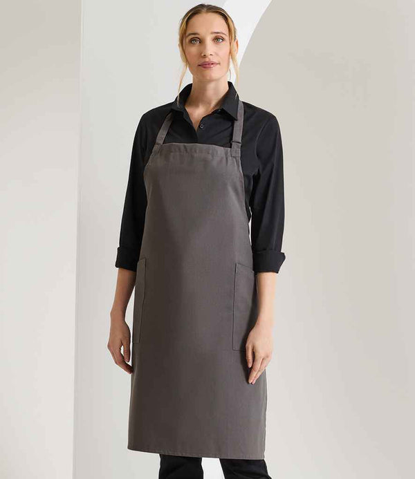 A woman faces forward wearing a black collared shirt and the Premier Recycled Polyester and Fairtrade Organic Cotton Bib Apron in grey, featuring front and side pockets. The background is plain with white and light grey walls.
