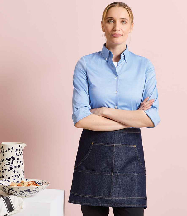 A person with blonde hair tied back, wearing a light blue easy care button-up shirt and the Premier Denim Waist Apron by Premier, stands with arms crossed in front of a pale pink background. A white table with a spotted pitcher and bowl is beside them.