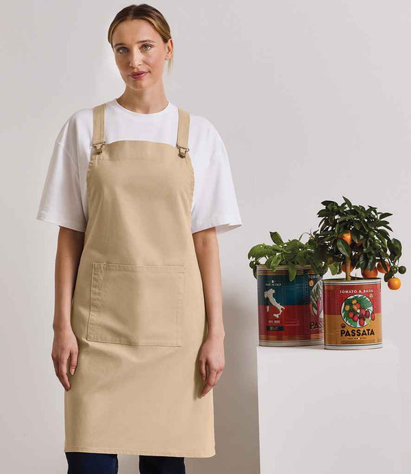 A person wearing the Premier Cross Back Barista Bib Apron by Premier over a white t-shirt stands next to a white table with a small potted plant and large tomato-labeled cans, against a plain, light-colored background.