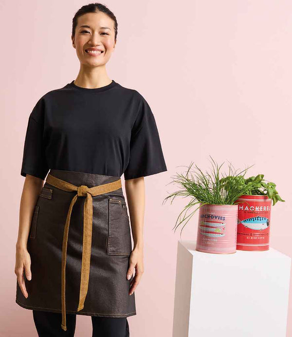 A person smiles by a white table, wearing a black t-shirt and the Premier Division Waist Apron by Premier, featuring a brown tie, holding two cans used as planters for green herbs against a light pink background.