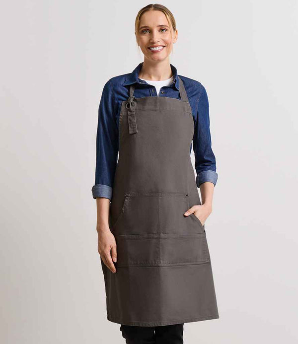A woman smiles at the camera, standing against a plain background in a blue rolled-sleeve shirt and the Premier Calibre Heavy Canvas Pocket Apron by Premier, with one hand resting in the aprons pocket.
