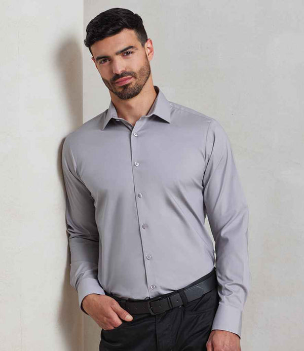 A man with short dark hair and a beard stands against a light wall, wearing the Premier Long Sleeve Stretch Fit Poplin Shirt by Premier in light gray and black pants with a black belt.