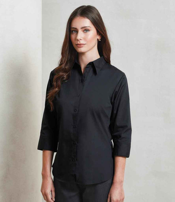 A person with long brown hair stands against a light wall, wearing the Premier Ladies 3/4 Sleeve Poplin Blouse by Premier in black, paired with black pants. They face forward with a neutral expression.