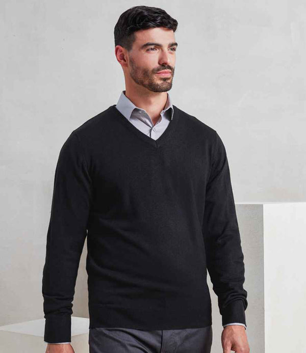 A man with short dark hair and a beard models the Premier Essential Acrylic V Neck Sweater by Premier in black, layered over a light gray collared shirt. He stands indoors against a plain, light-colored background, displaying modern style.