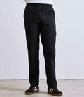 A person is wearing Premier Essential Chefs Cargo Trousers by Premier, featuring cargo pockets, an elasticated waistband, and a relaxed fit, paired with black sneakers with white stripes, standing indoors against a plain light background.