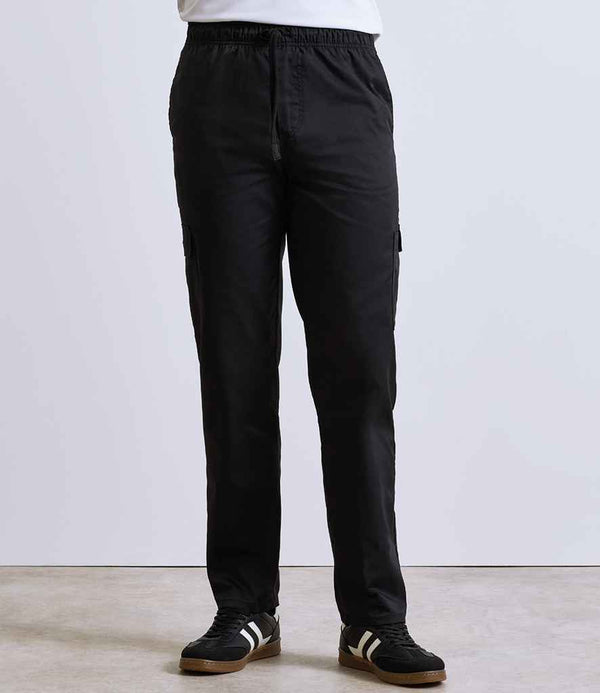 A person is wearing Premier Essential Chefs Cargo Trousers by Premier, featuring cargo pockets, an elasticated waistband, and a relaxed fit, paired with black sneakers with white stripes, standing indoors against a plain light background.