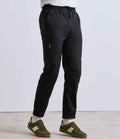A person stands against a plain background wearing Premier Recyclight® Chefs Cargo Trousers by Premier—black, lightweight, with an elasticated waistband, side zipper pocket, and tapered leg—paired with white socks and green-and-white sneakers.