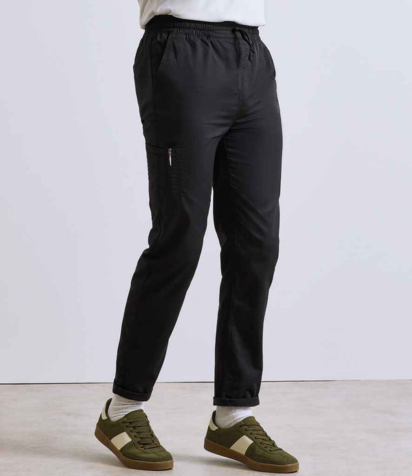 A person stands against a plain background wearing Premier Recyclight® Chefs Cargo Trousers by Premier—black, lightweight, with an elasticated waistband, side zipper pocket, and tapered leg—paired with white socks and green-and-white sneakers.