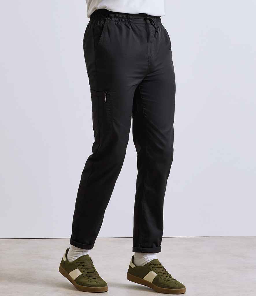 A person stands against a plain background wearing Premier Recyclight® Chefs Cargo Trousers by Premier—black, lightweight, with an elasticated waistband, side zipper pocket, and tapered leg—paired with white socks and green-and-white sneakers.