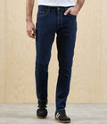 A person in Premier Workready Straight Leg Jeans by Premier, a white shirt, and black sneakers with white stripes stands against a light wood-paneled wall and gray floor, right hand in their pocket.