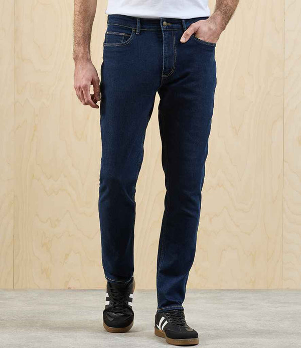 A person in Premier Workready Straight Leg Jeans by Premier, a white shirt, and black sneakers with white stripes stands against a light wood-paneled wall and gray floor, right hand in their pocket.