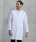 A bearded man with long hair tied back stands against a light gray concrete wall, smiling to his right in a Premier Essential 3 Pocket Work Coat by Premier over a white shirt and black pants.