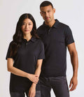 A woman and a man stand side by side against a plain light background, both wearing Premier Coolchecker® Stud Piqué Polo Shirts by Premier with dark pants.
