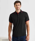 A man is wearing the Premier Contrast Coolchecker® Piqué Polo Shirt by Premier, featuring a ribbed collar with red trim on the sleeves, paired with blue jeans. He stands facing forward and looks at the camera against a plain background.