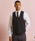 A man stands against a light pink background, wearing the Premier Lined Waistcoat by Premier over a white shirt and black tie, paired with dark pants. He faces the camera with one hand behind his back.