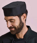 A bearded man wears a black Premier Turn-Up Chefs Hat and a black, easy-care chef’s jacket. He looks down with a slight smile against a light pink background.