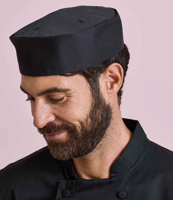 A bearded man wears a black Premier Turn-Up Chefs Hat and a black, easy-care chef’s jacket. He looks down with a slight smile against a light pink background.