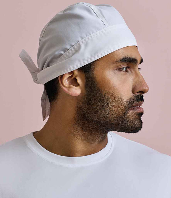 A man with a short beard and mustache, wearing a Premier Chefs Zandana by Premier and an easy care white shirt, looks to the right against a plain light pink background.