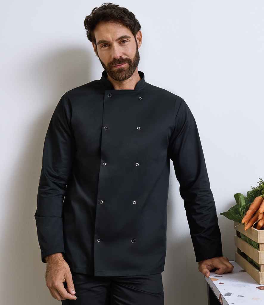 A man with dark hair and a beard wears the Premier Unisex Long Sleeve Stud Front Chefs Jacket by Premier, standing indoors with one hand on a table holding a crate of carrots and leafy greens, facing the camera against a plain white wall.