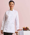 A woman wears the Premier Ladies Long Sleeve Chefs Jacket by Premier, standing beside a table with assorted macarons and strawberries against a plain pink background.