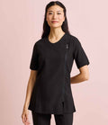 A woman with long dark hair stands against a light pink background, wearing the Premier Ladies Camellia Short Sleeve Tunic by Premier, featuring an asymmetric front zip and easy-care fabric for modern comfort.