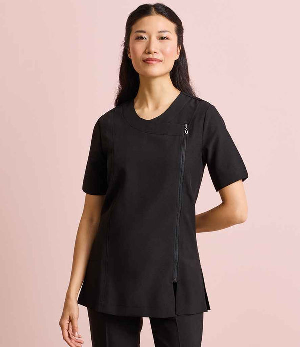 A woman with long dark hair stands against a light pink background, wearing the Premier Ladies Camellia Short Sleeve Tunic by Premier, featuring an asymmetric front zip and easy-care fabric for modern comfort.