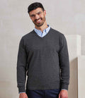 A man with short dark hair and a beard is standing and smiling. He is wearing a light blue shirt under the Premier Knitted Cotton Acrylic V Neck Sweater by Premier, paired with dark pants, in front of a plain light background.