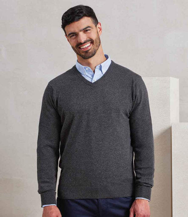 A man with short dark hair and a beard is standing and smiling. He is wearing a light blue shirt under the Premier Knitted Cotton Acrylic V Neck Sweater by Premier, paired with dark pants, in front of a plain light background.