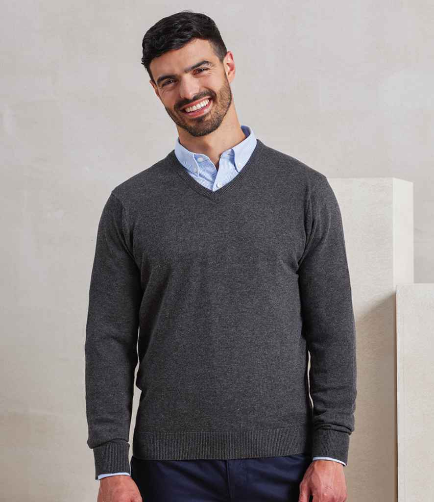 A man with short dark hair and a beard is standing and smiling. He is wearing a light blue shirt under the Premier Knitted Cotton Acrylic V Neck Sweater by Premier, paired with dark pants, in front of a plain light background.