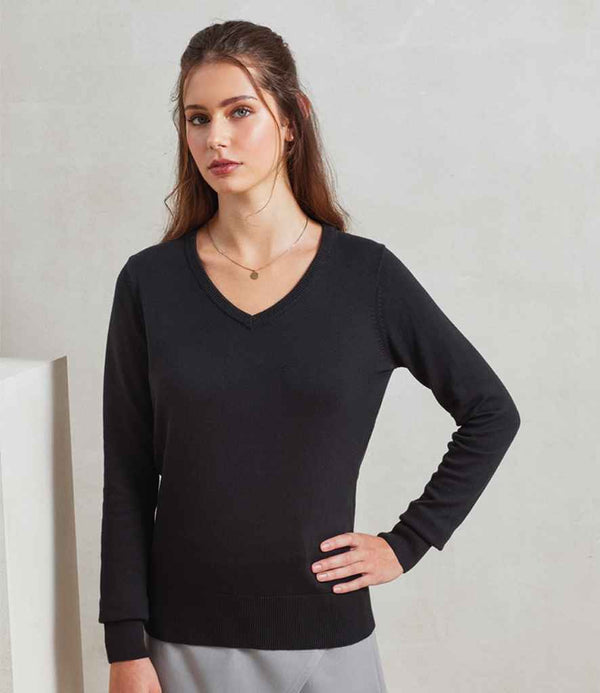 A woman with long brown hair wears the Premier Ladies Knitted Cotton Acrylic V Neck Sweater in black, paired with light gray pants. She stands indoors, one hand on her hip, against a plain light background.