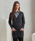 A woman with long brown hair stands indoors, hands in her pockets, wearing the Premier Ladies Longline V Neck Cardigan by Premier over a gray collared shirt and black pants against a plain, neutral background.