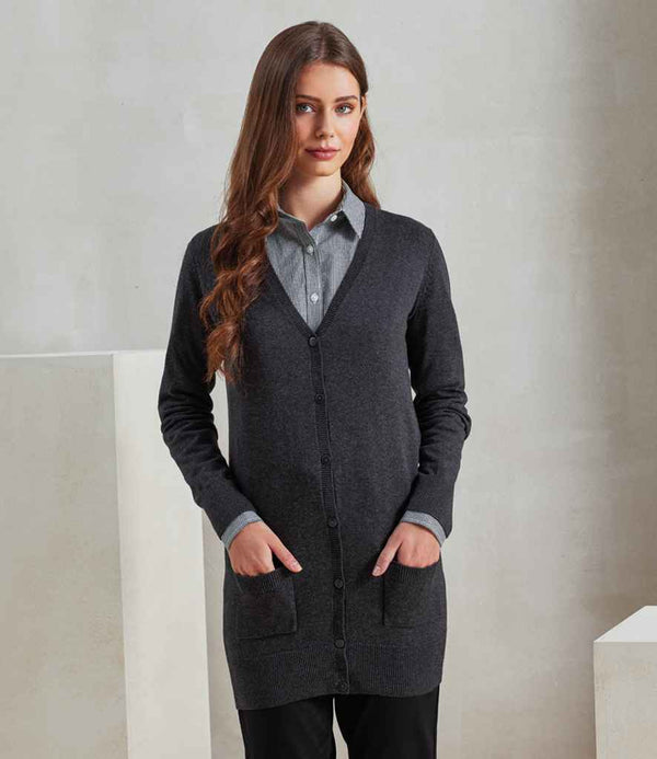 A woman with long brown hair stands indoors, hands in her pockets, wearing the Premier Ladies Longline V Neck Cardigan by Premier over a gray collared shirt and black pants against a plain, neutral background.